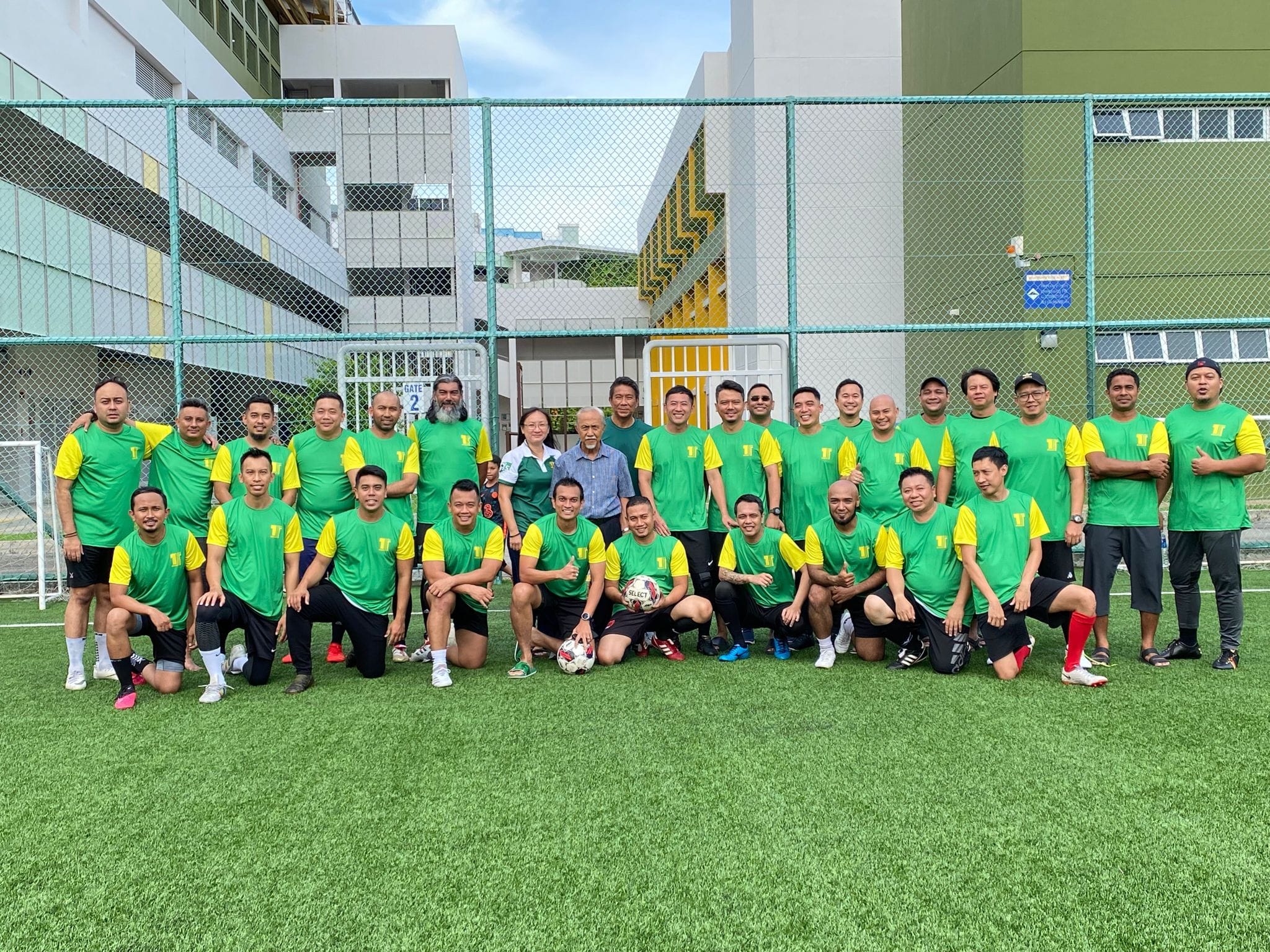 A large group of men in green and yellow jerseys pose on an outdoor football pitch with a soccer ball.